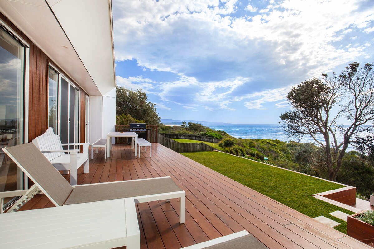A spacious deck offers lounge chairs for relaxation, facing the ocean and showcasing a view of the Tasman Sea. A BBQ area is visible, surrounded by green grass and coastal vegetation under a partly cloudy sky.