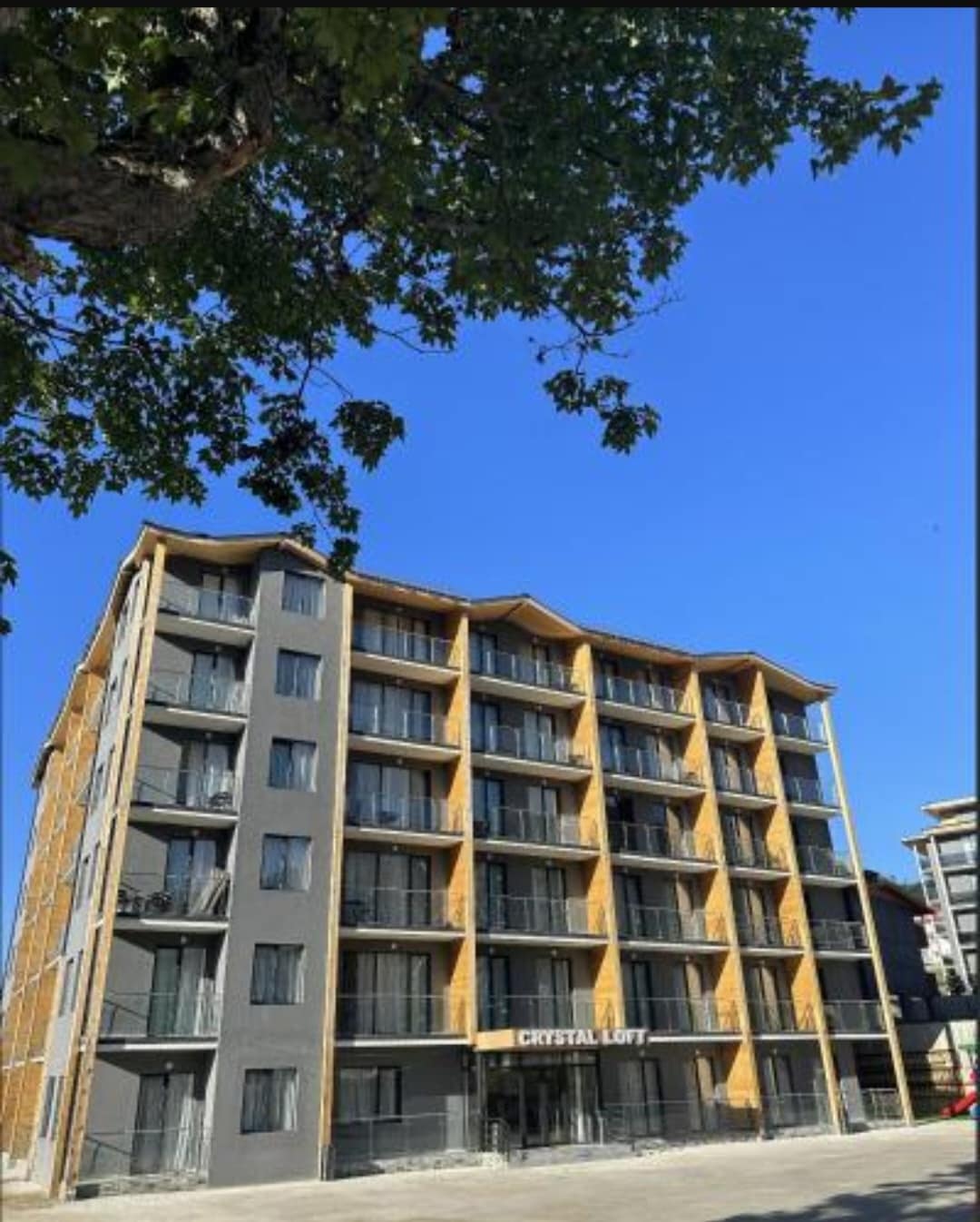 The exterior of the Crystal Loft building is showcased, featuring a modern design with a mix of wood and concrete. Several balconies can be seen on the front of the structure, and bright blue skies create a vivid backdrop.