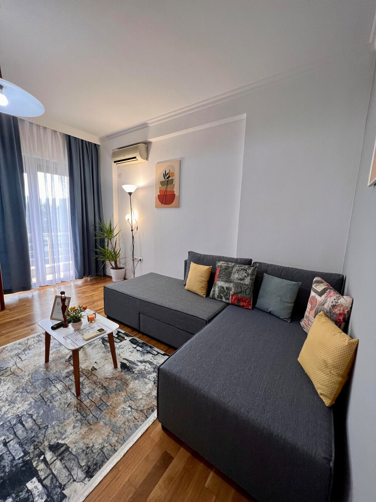 A comfortable living area features a gray sectional sofa adorned with colorful cushions. A small coffee table is positioned in front of the sofa, resting on a patterned rug. Natural light floods the space through large glass doors that lead to a balcony.