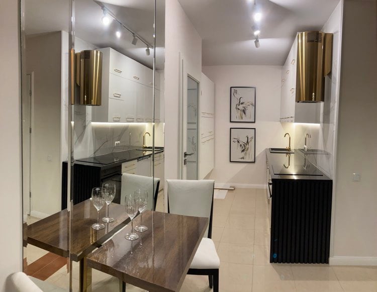 A modern kitchen area features sleek cabinetry with gold accents and a contrasting black countertop. A small dining table is set for two, complemented by white chairs. Minimalist wall art enhances the neutral color palette, and soft lighting creates a welcoming atmosphere.