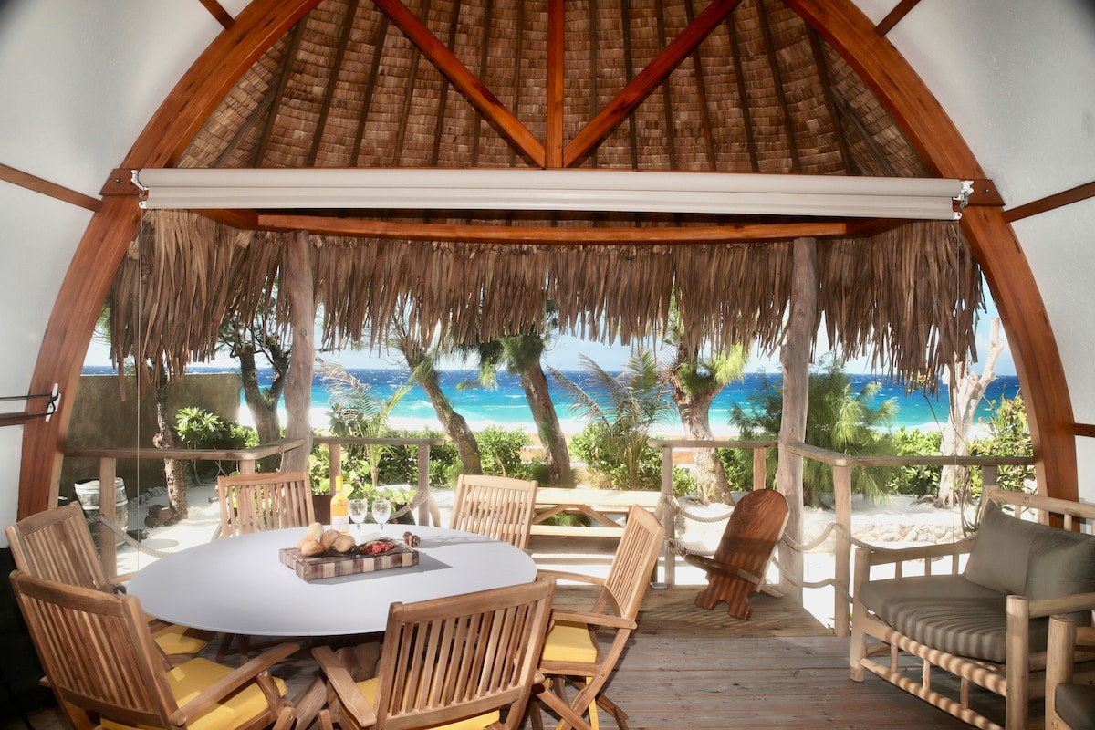 A spacious outdoor seating area is highlighted with a round table surrounded by wooden chairs and a comfortable lounge chair. A thatched roof provides shade, while views of the beach and ocean are visible through open walls, inviting a natural breeze.