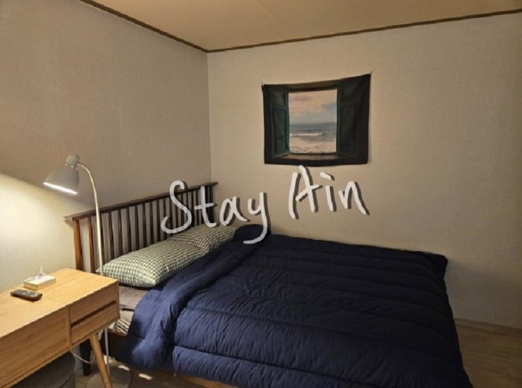 A calming bedroom features a spacious bed covered with a dark blue comforter. A bedside table holds a lamp and a small device, while a window with a scenic view is adorned with a dark curtain. Neutral walls create a serene atmosphere.