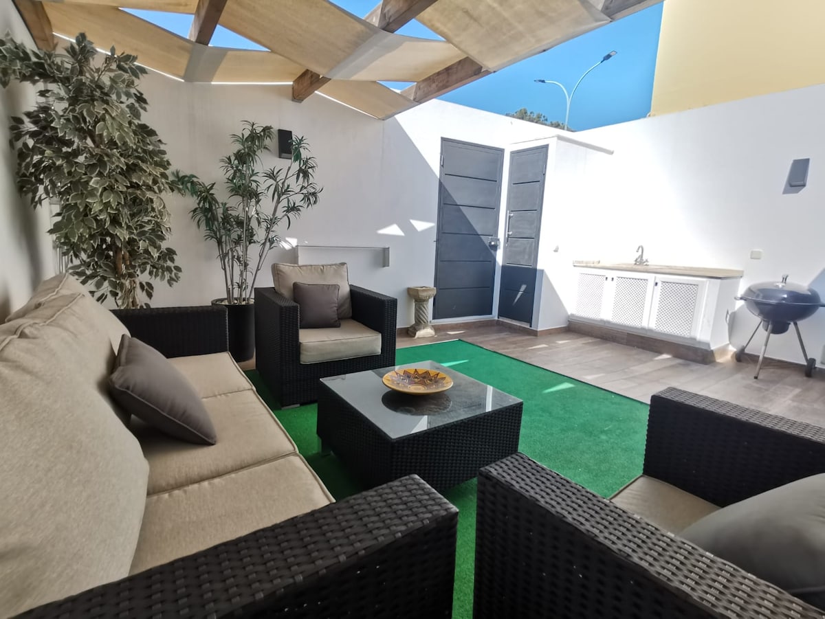 A bright outdoor patio space is equipped with comfortable seating on dark wicker furniture. A shaded area is created by a simple canopy, while an outdoor kitchen setup and a barbecue grill are present. Green turf flooring adds a fresh touch to the area.
