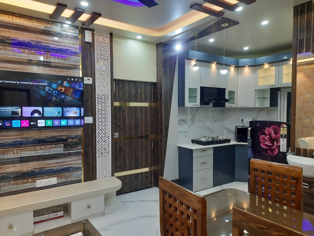 A modern living space features a large flat-screen TV mounted on a decorative wall, paired with a sleek dining table. The kitchen area is outfitted with new appliances, including a black microwave and a gas stove, complemented by contemporary cabinetry.