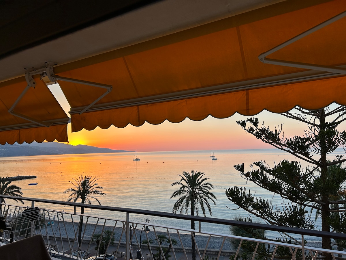 A vibrant sunset is seen over calm waters, with silhouettes of palm trees framing the scene. Soft orange and pink hues fill the sky, while sailboats rest peacefully on the horizon, creating a serene coastal atmosphere.