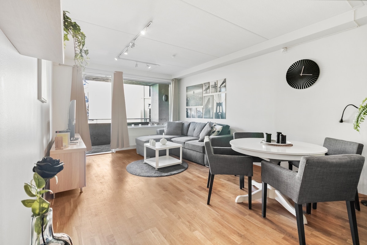 A spacious living area is highlighted by a large gray sofa and a round dining table surrounded by four chairs. Natural light floods in from a large window, alongside decorative wall art. A private balcony is visible through the glass doors, enhancing the open feel.