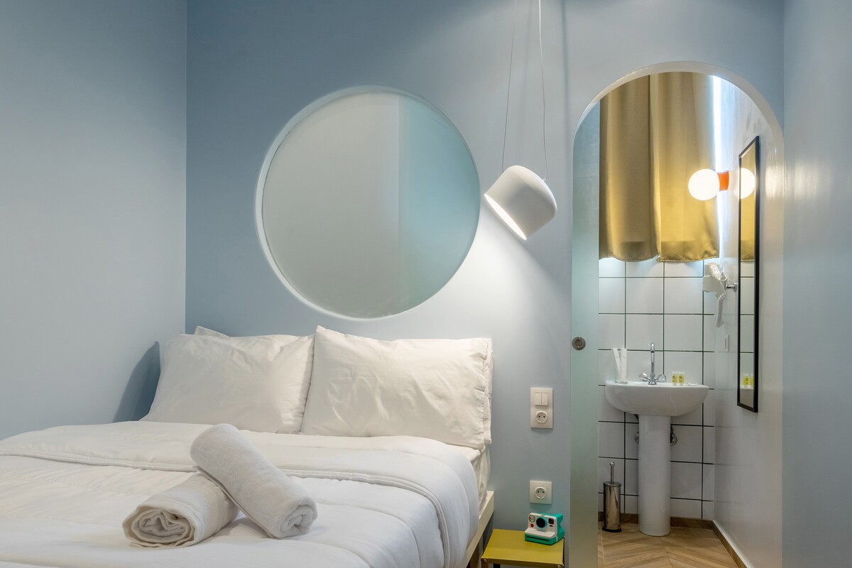 A well-lit room features a spacious bed with crisp white linens and fresh towels arranged neatly. A circular mirror and pendant light enhance the design. A compact bathroom area is visible, showcasing modern fixtures with white tiles and a stylish sink.