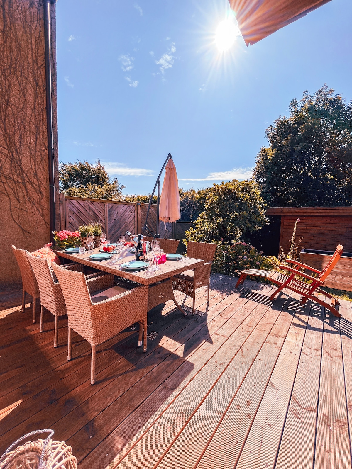 An outdoor dining area features a wooden table surrounded by six wicker chairs, set against a backdrop of lush greenery. A sun umbrella is positioned to one side, and a wooden lounge chair rests nearby. Bright sunlight illuminates the entire space.