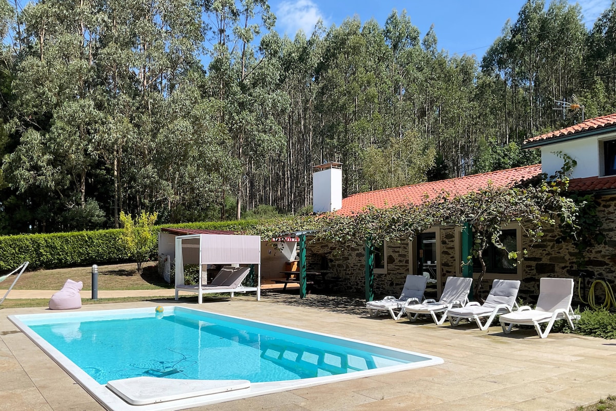 A clear blue swimming pool is framed by a spacious stone patio, featuring several white lounge chairs arranged along one side. Behind the pool, a charming rustic building is partially covered in greenery, with tall trees providing a serene backdrop.