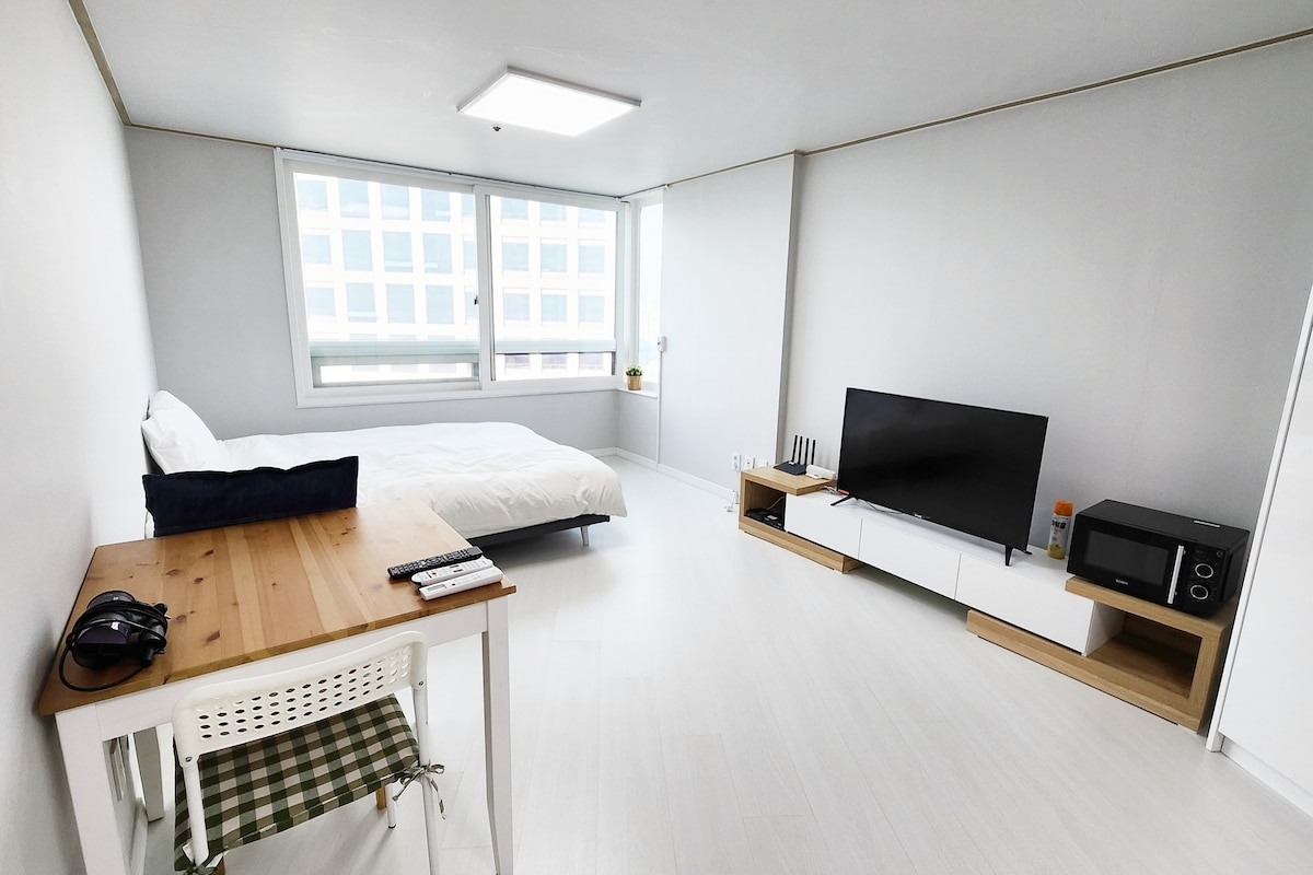 A bright and airy room features a spacious bed with white bedding and a large window allowing natural light. A wooden table with a checkered chair is positioned next to a television on a minimalist stand, while a microwave is subtly placed on a shelf.