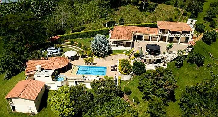 An aerial view of the property showcases a spacious layout surrounded by greenery. A clear blue pool is visible alongside several lounging areas, with a main building featuring multiple balconies and terracotta rooftops. Nearby, fruit trees and manicured gardens enhance the natural environment.