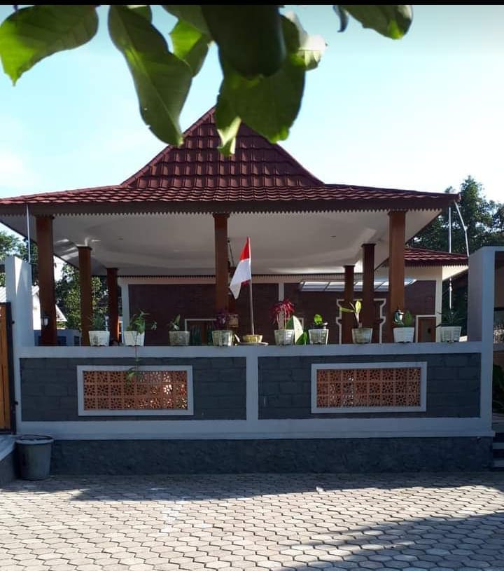 A traditional gazebo with a red tiled roof is situated within a landscaped area. Potted plants adorn the gazebo's base, and a national flag is displayed prominently. The surrounding wall features decorative lattice work, and the ground is paved with stone.