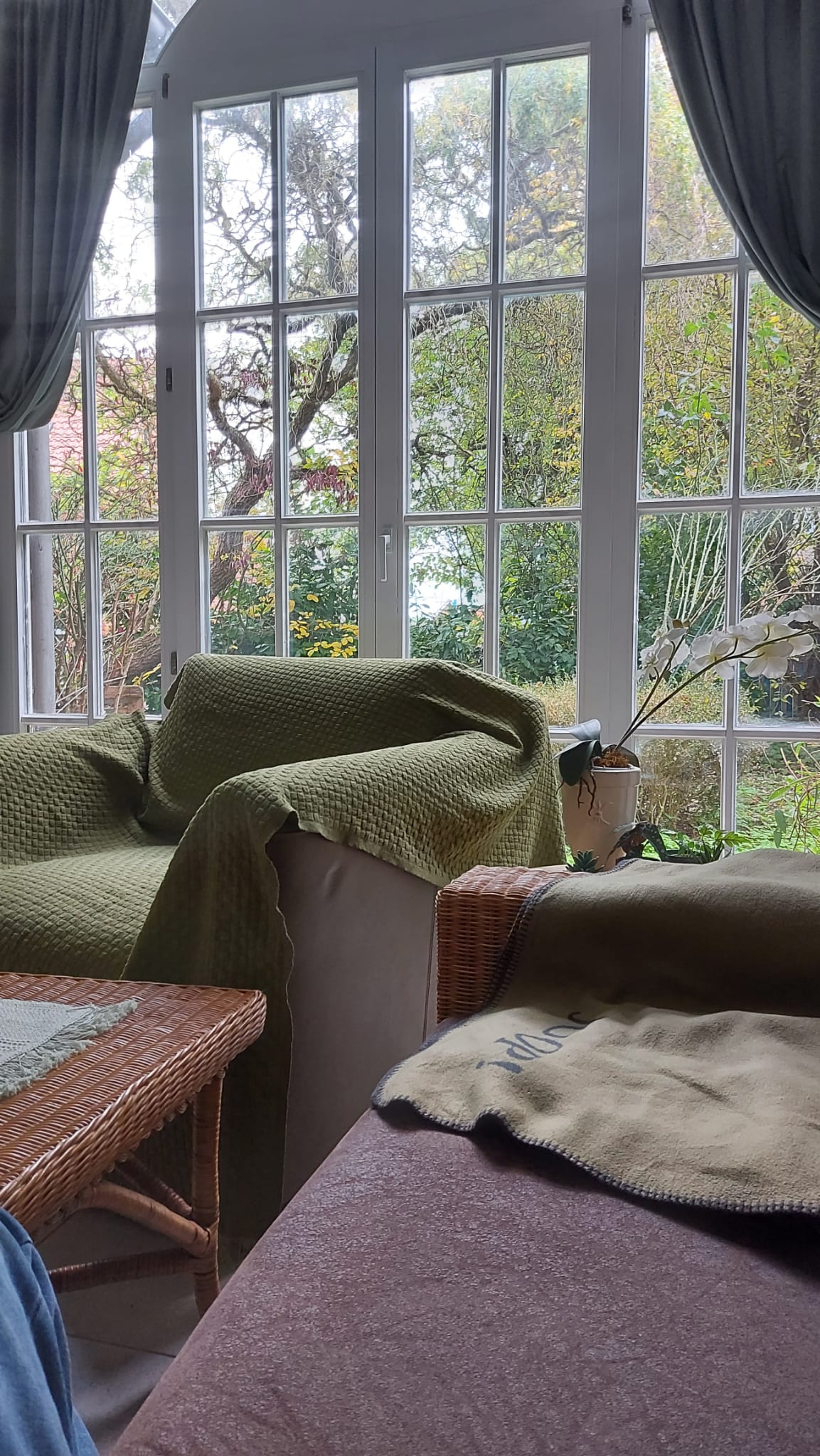 A cozy living area is featured, with two armchairs draped in soft blankets. Large windows adorned with flowing curtains showcase a lush garden view, allowing natural light to brighten the space. A small table adds functionality next to the seating arrangement.
