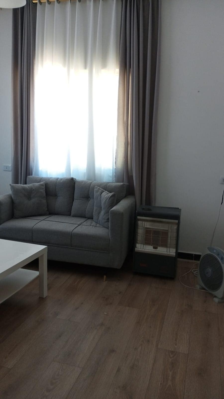A comfortable gray sofa with plush cushions is positioned against a wall beneath soft white curtains. A small white coffee table is placed in front of the sofa, and a fan can be seen beside a heater, all situated on warm-toned wooden flooring.