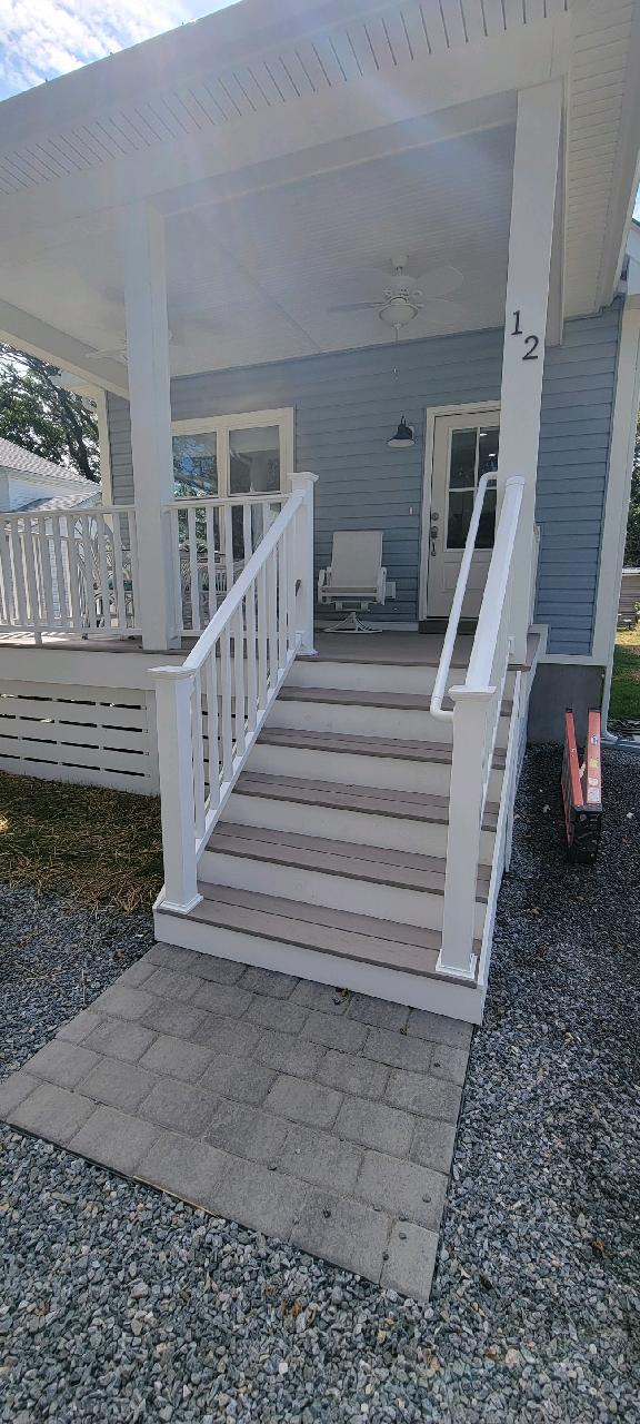 A welcoming porch is depicted, featuring a set of white steps leading up to a light blue house. A rocking chair sits to the side, with a small light fixture hanging nearby. Pebbled pathways and well-kept landscaping frame the entrance.