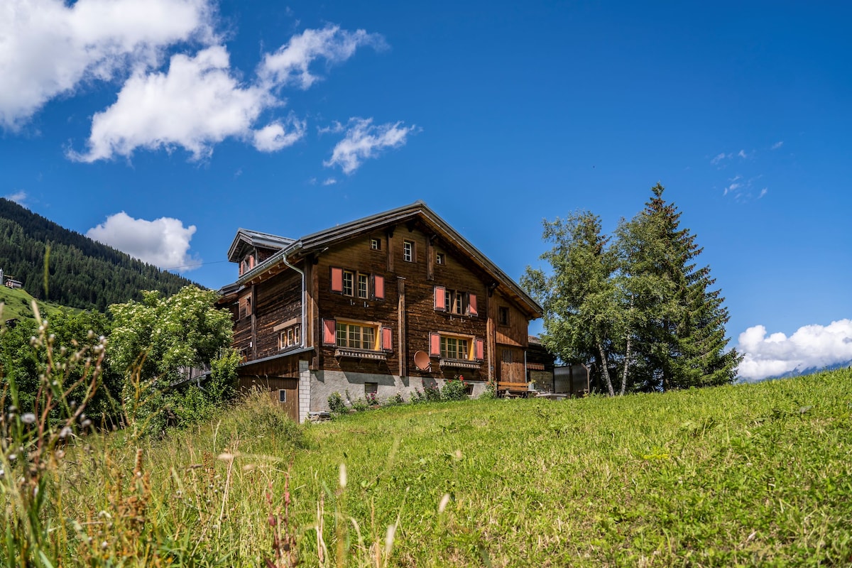 A traditional wooden chalet is set against a clear blue sky, surrounded by lush greenery. The façade features pink shutters, and large windows reflect the natural landscape. A gentle slope leads to the entrance, enhancing the home's serene environment.