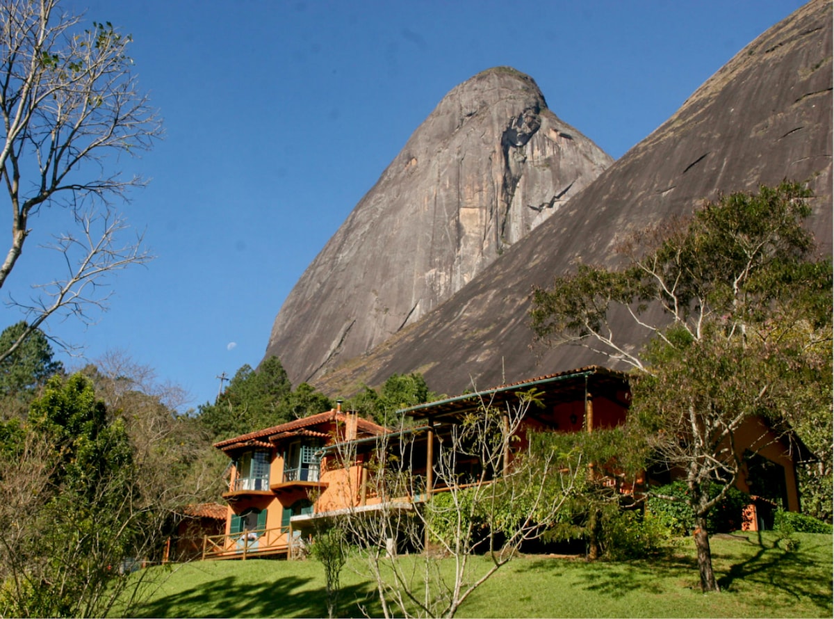 The exterior of a multi-level house is nestled against a dramatic mountain backdrop. Lush greenery surrounds the property, contributing to a tranquil environment. Large windows invite natural light, and a wooden deck is visible in front, offering a connection to the serene landscape.
