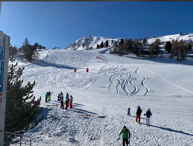 A wide view of a snowy ski slope features various ski tracks made by past skiers. Groups of individuals are seen enjoying the snow, with a mix of adults and children. The backdrop showcases snow-covered mountains under a clear blue sky, creating a serene winter environment.