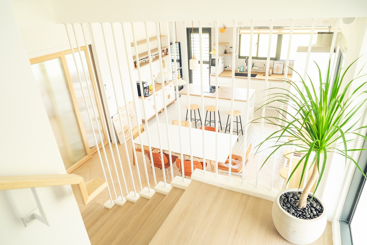 A bright and open living space is visible from an upper level. The area features a modern kitchen with light wood cabinetry and bar stools, alongside a dining area with spacious tables. A decorative plant adds a touch of greenery to the setting.