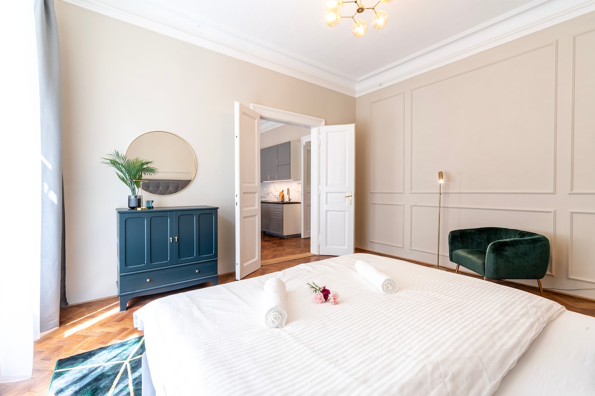 A spacious bedroom features a large bed dressed in crisp white linens, with two rolled towels placed neatly on top. A green velvet chair adds a touch of elegance, and a round mirror is positioned above a stylish blue cabinet. Natural light streams through sheer curtains.
