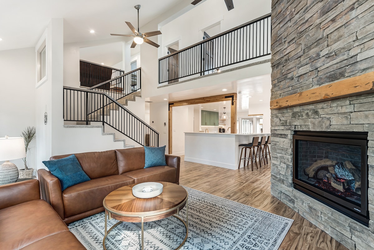 A spacious living area features a rich leather sectional sofa, complemented by a circular coffee table and decorative accents. Nearby, a stone fireplace adds warmth, while a staircase leads to the upper level overlooking the open floor plan, creating a welcoming ambiance.