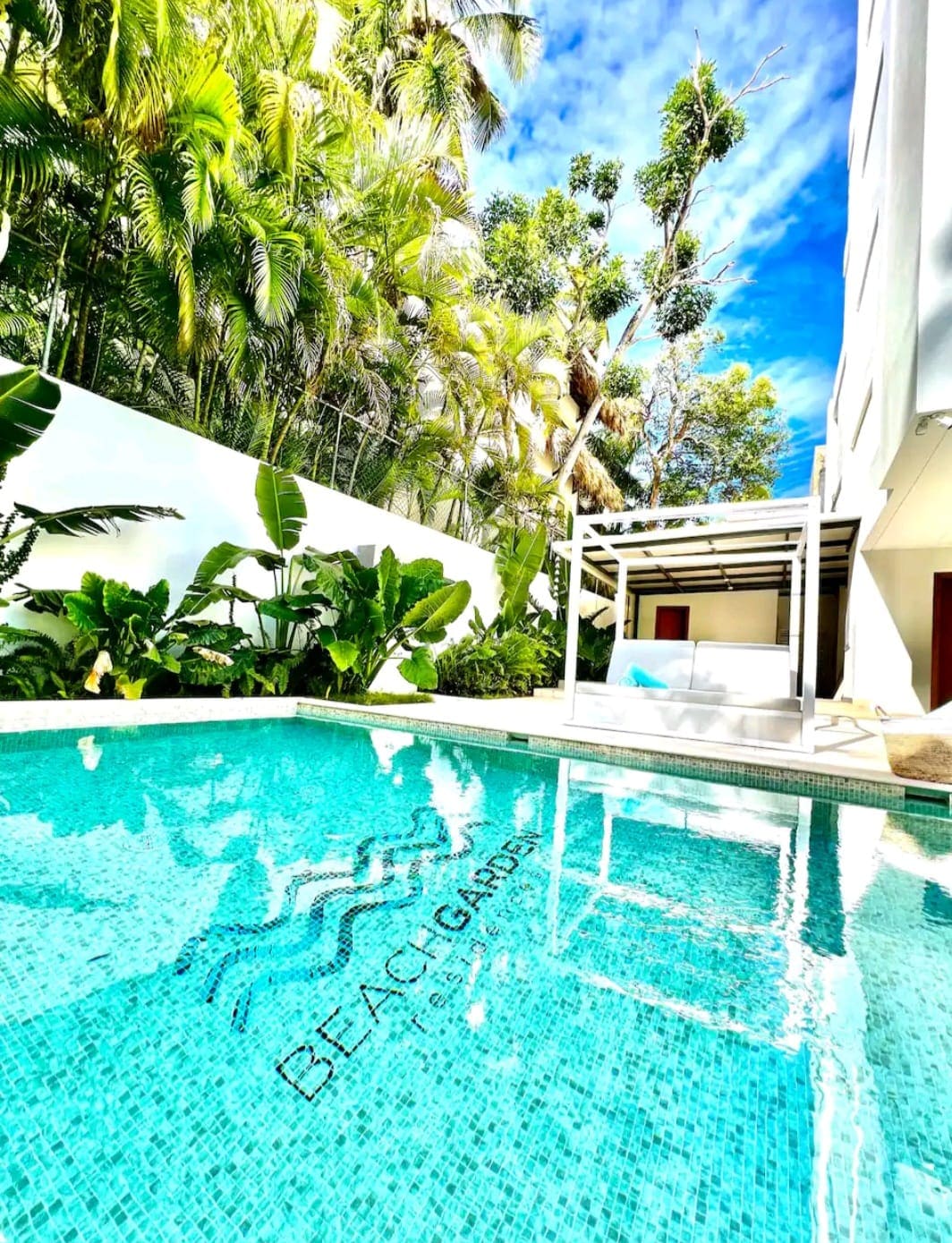 A private pool is depicted, featuring clear turquoise water that reflects the blue sky above. Lush green foliage surrounds the area, enhancing the sense of privacy. A sheltered seating area is visible beside the pool, providing a serene space for relaxation.
