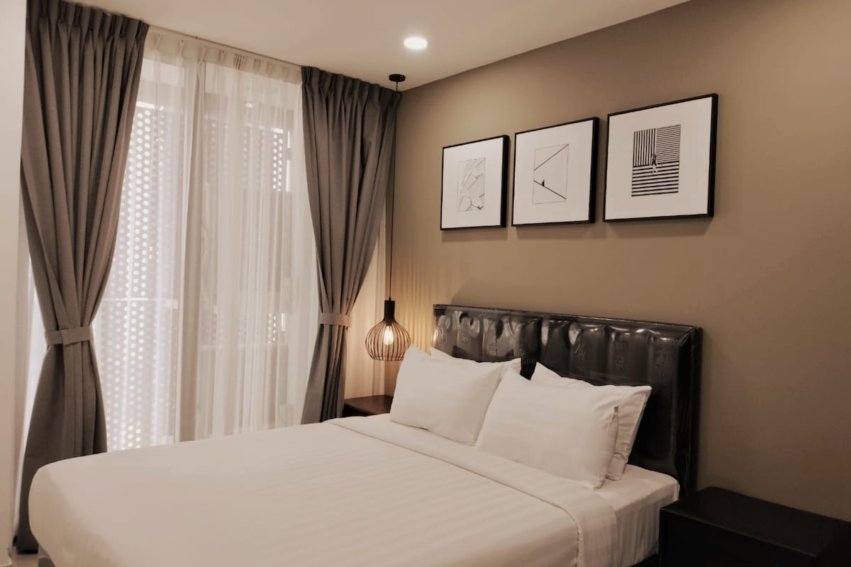 A cozy sleeping area features a spacious bed with crisp white bedding against a muted wall. Stylish wall art hangs above the headboard. Soft, sheer curtains allow natural light to filter through, complemented by elegant bedside lighting.
