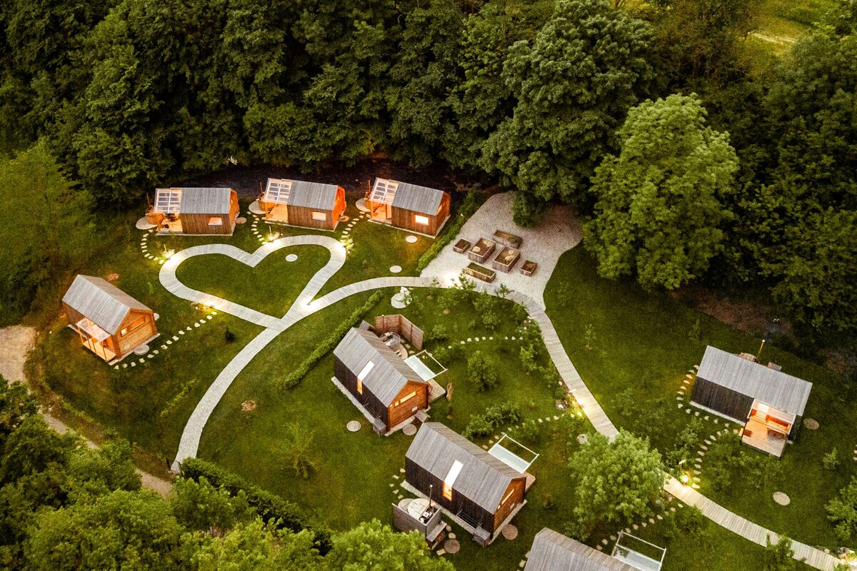 An aerial view of the glamping site reveals several wooden cabins arranged along winding paths. The area is surrounded by dense greenery, with warm lights outlining the heart shape on the ground, creating a welcoming ambiance. Lounge areas with seating can be seen intermittently.