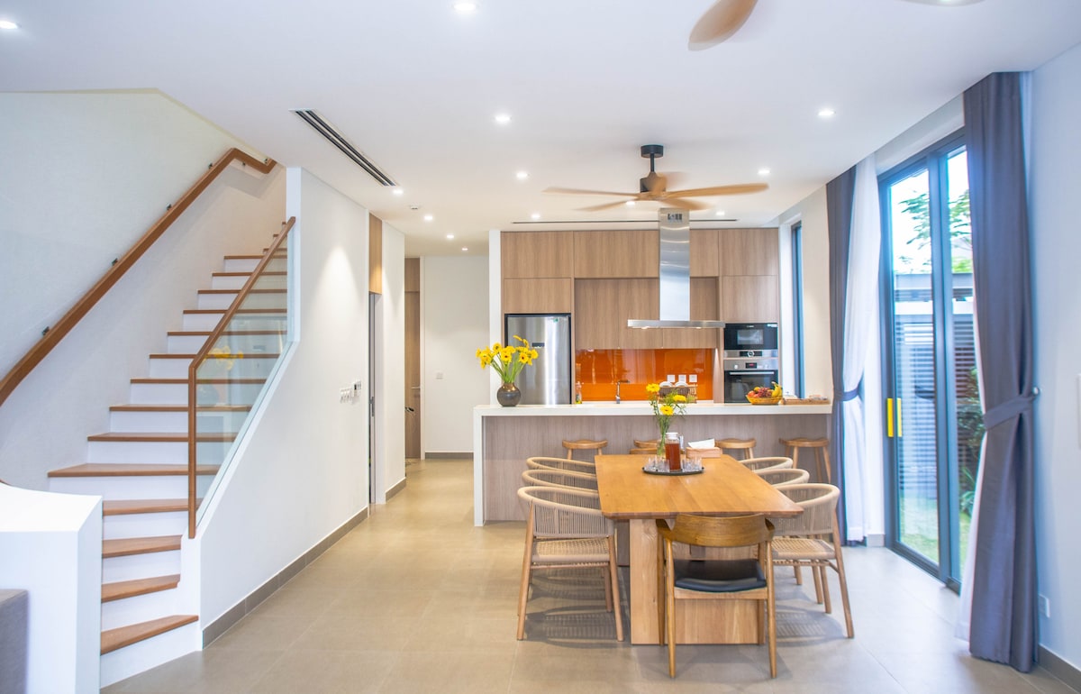 A modern dining area is enhanced by a large wooden table surrounded by wicker chairs. A sleek kitchen features a combination of upper and lower cabinets with stainless steel appliances, and tall glass doors lead to an outdoor space, allowing ample natural light.