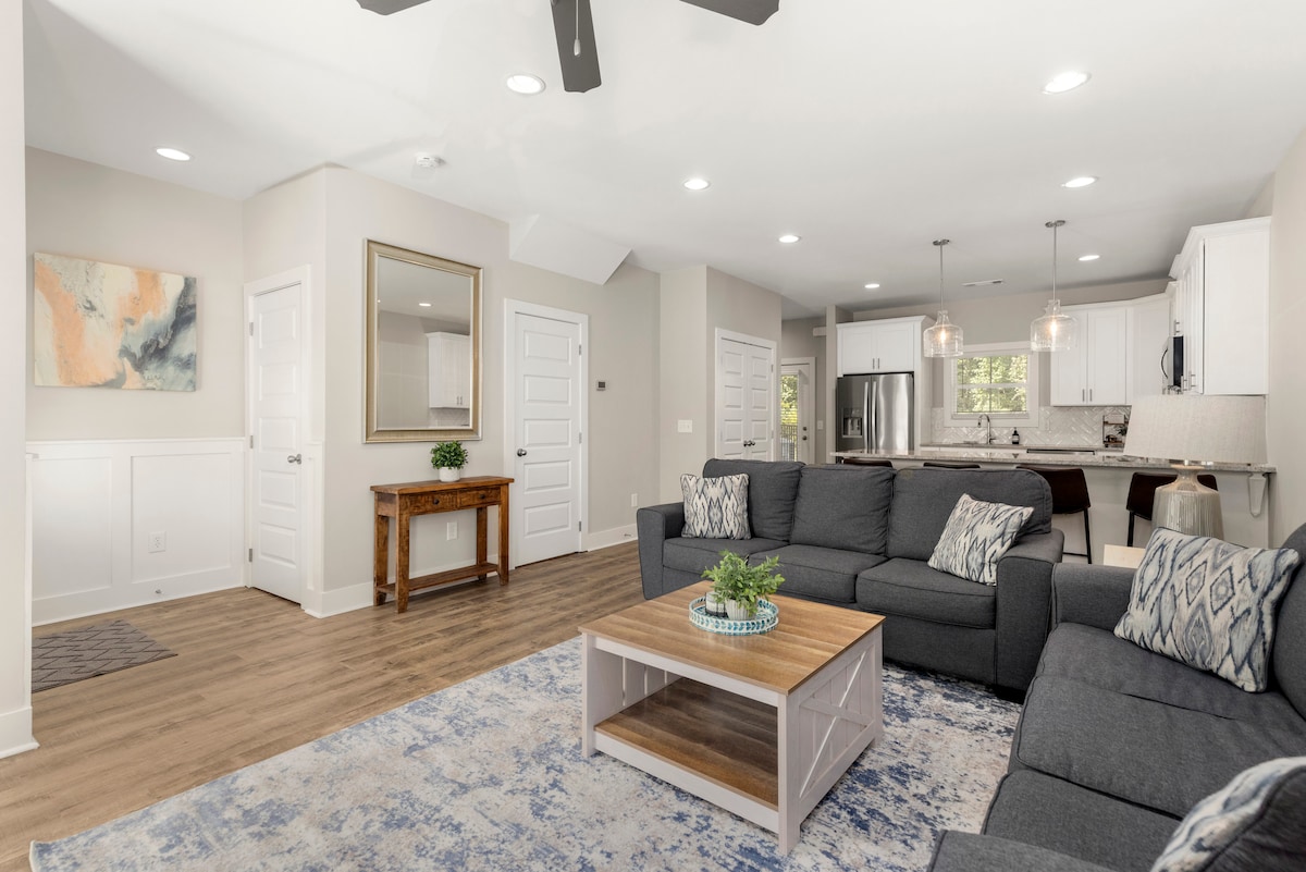 A spacious living area features a comfortable sectional sofa adorned with decorative pillows, facing a coffee table. Natural light illuminates the room, highlighting a neutral color palette. An open kitchen can be seen in the background, showcasing modern appliances and a dining area.