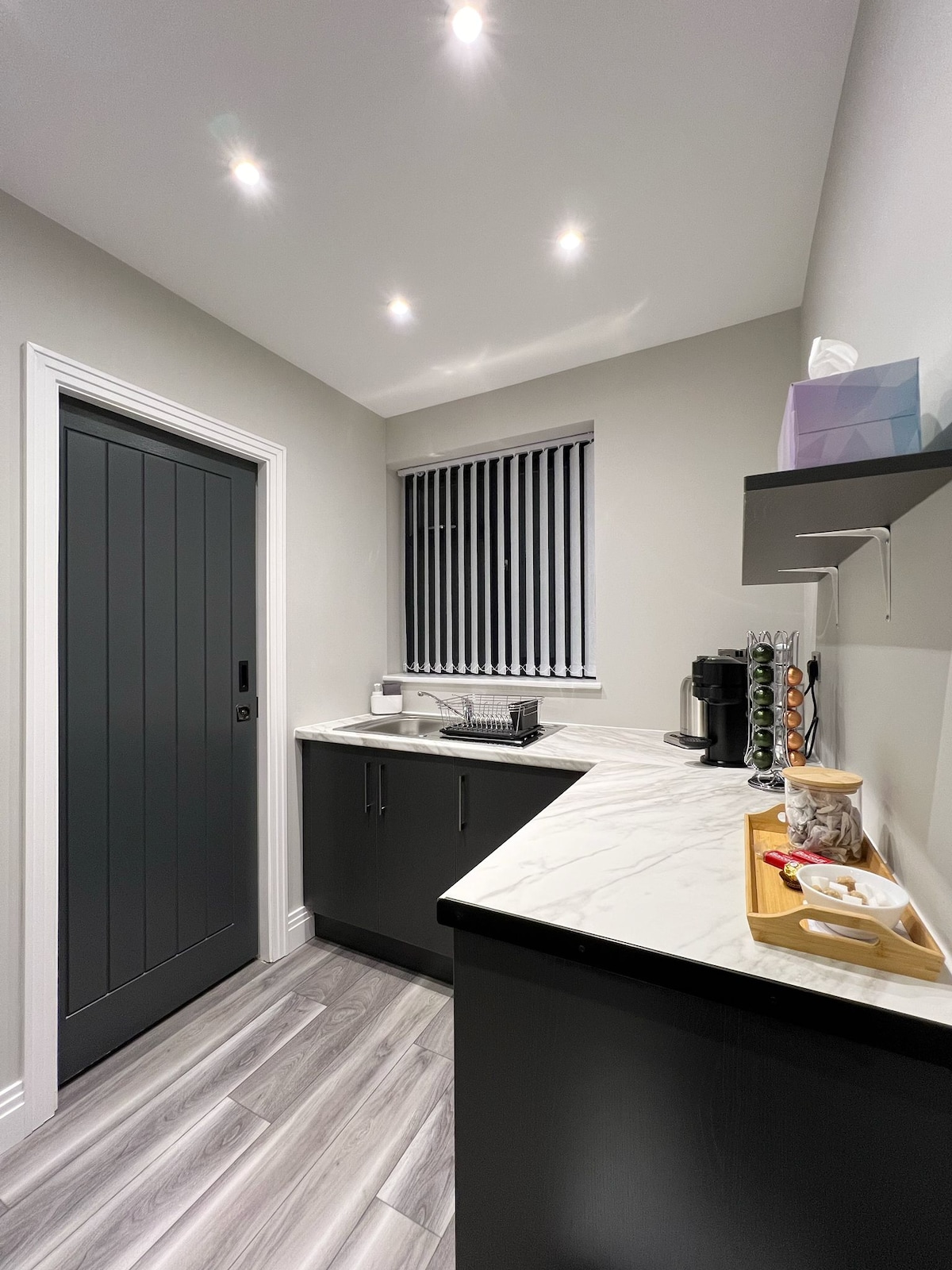 A compact kitchen features sleek dark cabinetry and a marble countertop. A window with blinds allows natural light, highlighting modern appliances including a coffee maker. Organized utensils and snacks are neatly arranged on a wooden tray, creating a practical cooking space.