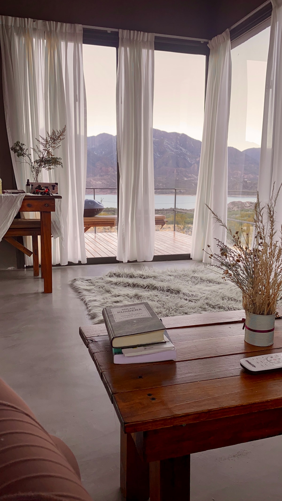 A living area is captured with large glass doors offering expansive mountain and lake views. Light, sheer curtains frame the windows. A wooden table is set nearby, accompanied by a cozy rug and potted plants, creating a peaceful and inviting atmosphere.
