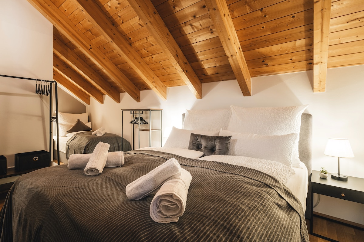 A cozy bedroom features two spacious beds with neutral bedding accentuated by neatly arranged towels. The wooden ceiling beams add warmth, while a small nightstand with a lamp provides soft illumination. A clothing rack is situated nearby, ensuring convenient storage space.