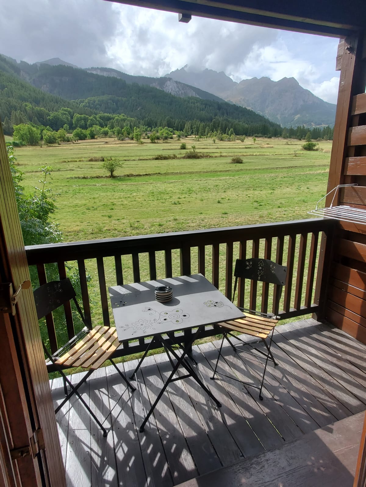 A balcony area is shown, featuring a small black table with two folding chairs. The surrounding landscape displays lush greenery and rolling hills, framed by distant mountains under a partly cloudy sky.