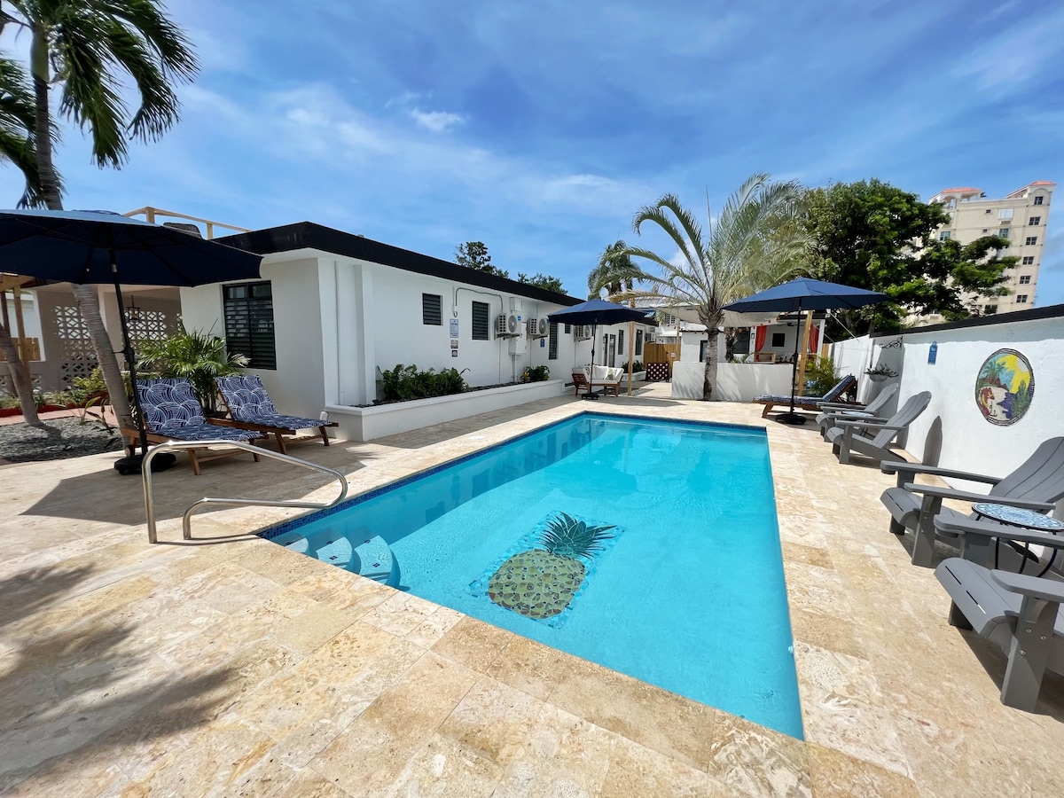 A clear blue pool reflects the sky, surrounded by lounge chairs and umbrellas for shade. Tropical plants frame the area, and a mural can be seen on the nearby wall. The suite entrance is situated in the background, inviting relaxation.