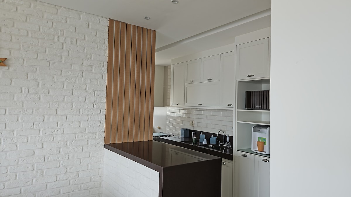 A modern kitchen area is visible, featuring white cabinetry with wooden accents. A dark counter contrasts with white brick walls. Essential appliances, including a microwave and coffee maker, are neatly arranged on the shelves, providing an organized space for meal preparation.