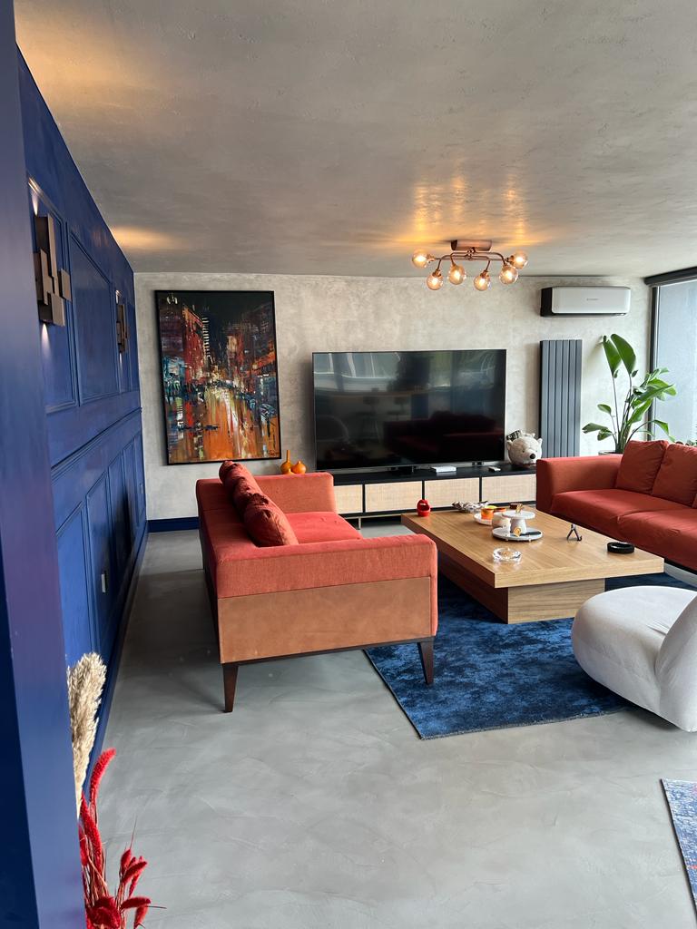 A modern living room features a large television against a textured wall, complemented by a rich blue accent wall. A spacious orange sofa and a coffee table with decorative items create a welcoming atmosphere. Large windows provide natural light and a view of the outdoor area.