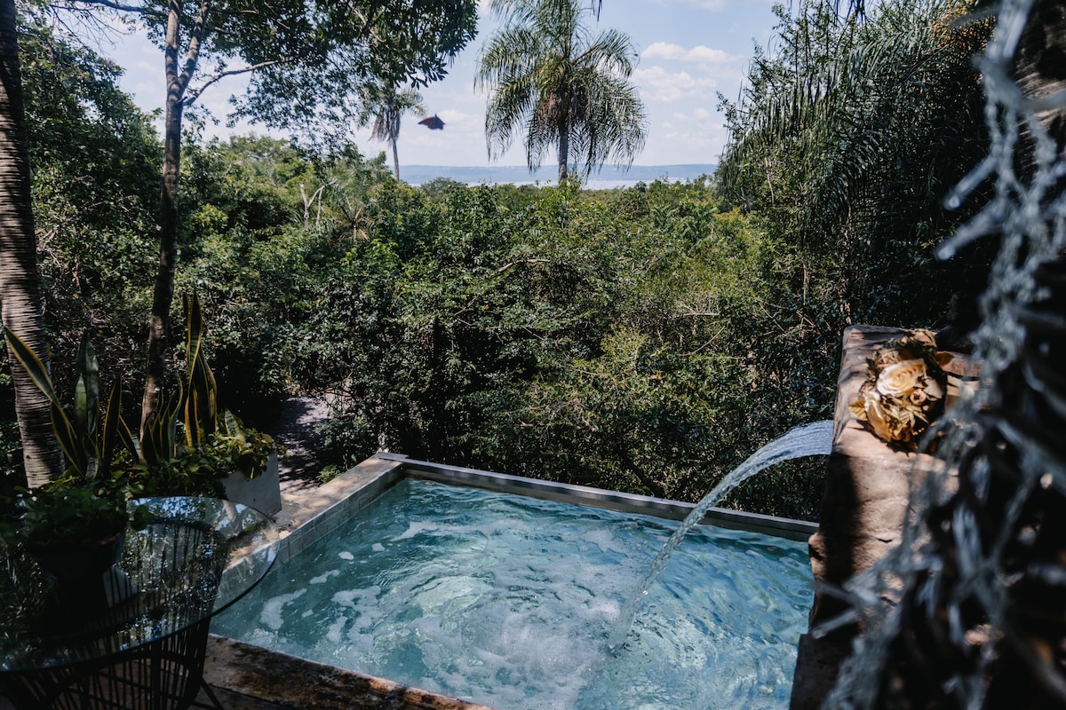 A serene plunge pool is framed by lush greenery, featuring a gentle waterfall effect. Sunlight filters through the surrounding trees, casting dappled shadows across the water's surface. In the background, distant views of the natural landscape can be seen, enhancing the tranquil setting.