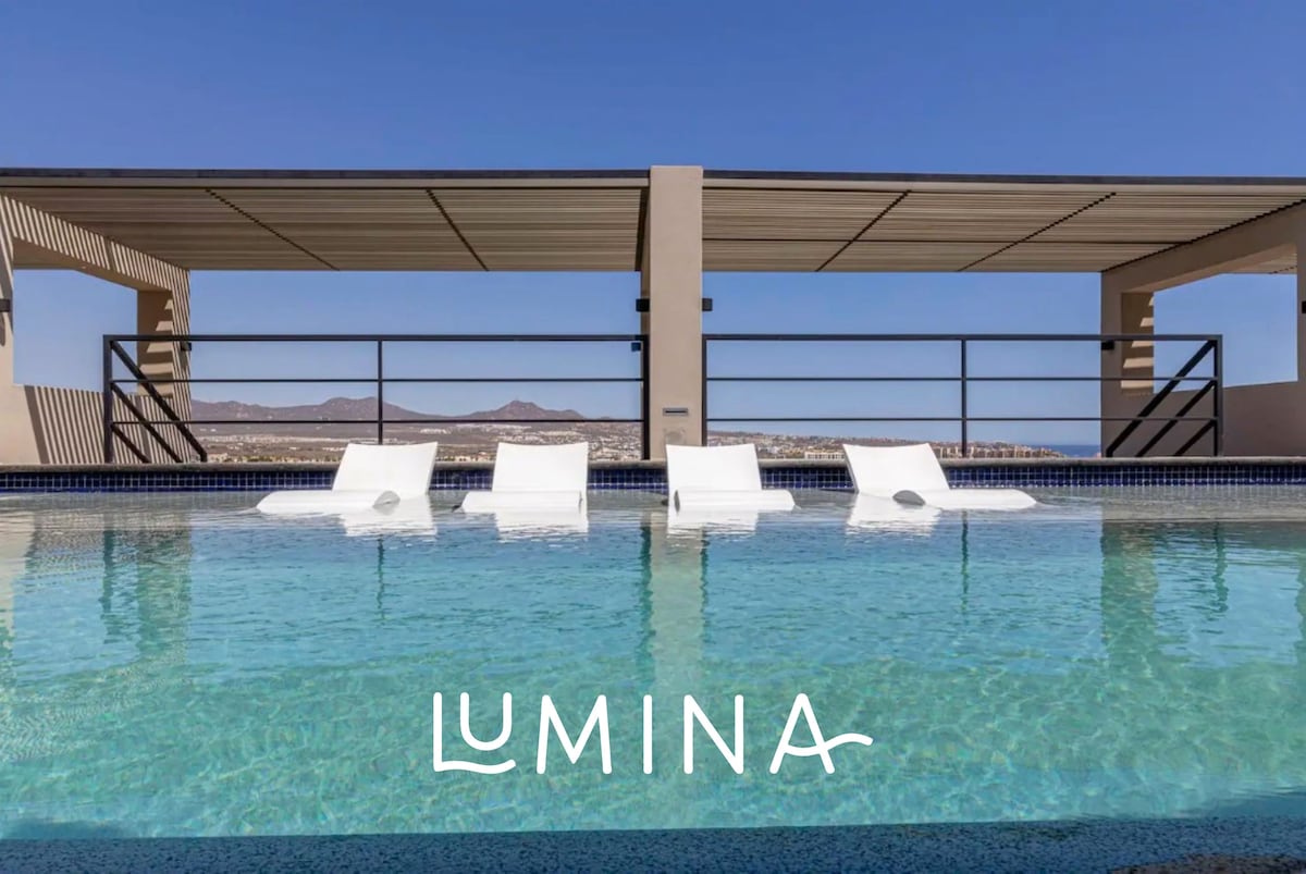 A serene rooftop pool area is shown, featuring white lounge chairs positioned partially in the water. Clear skies and distant mountains provide a backdrop, offering a tranquil space for relaxation.