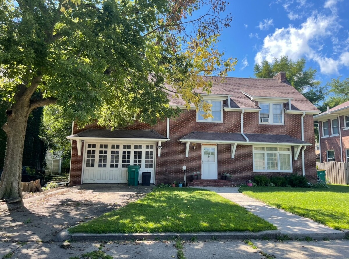 The exterior of a historic brick house is highlighted by a well-maintained lawn. Tall trees provide shade. The home features a covered entrance and a garage with multiple doors. A sidewalk leads to the entrance, enhancing the welcoming presence of the property.