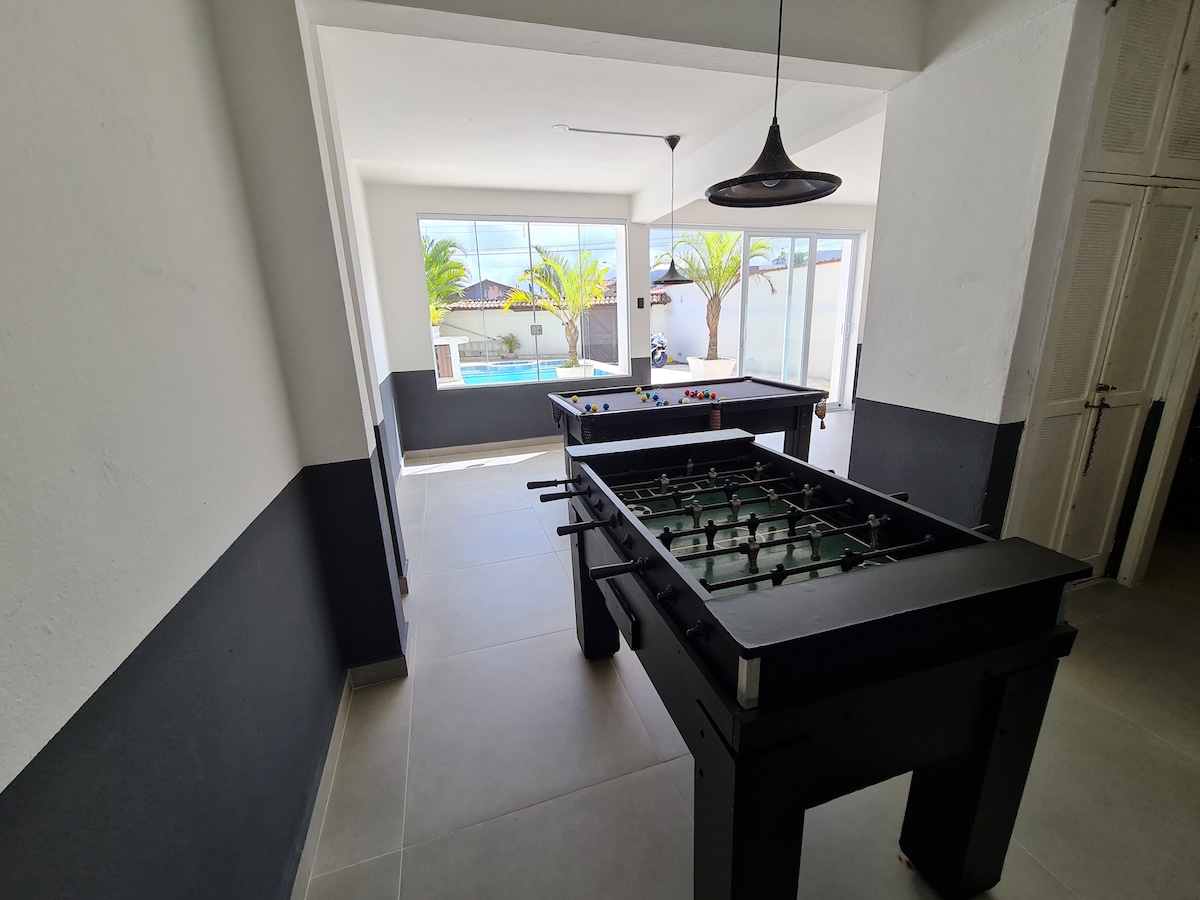 A game room is presented, featuring a foosball table and a pool table. Large windows allow natural light to fill the space, revealing views of the pool and palm trees outside. The walls are a two-tone design, with clean lines and a modern aesthetic.