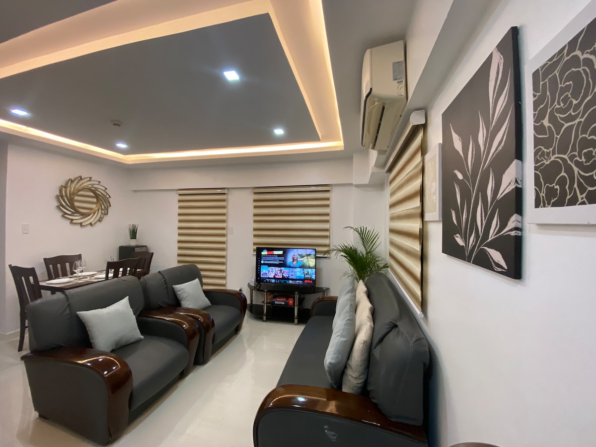 A spacious living area features modern furnishings with two comfortable sofas and a large flat-screen TV. Soft lighting from the ceiling highlights the decor, while natural light filters through dual-tone roller blinds. A dining area is visible in the background, accommodating family meals.