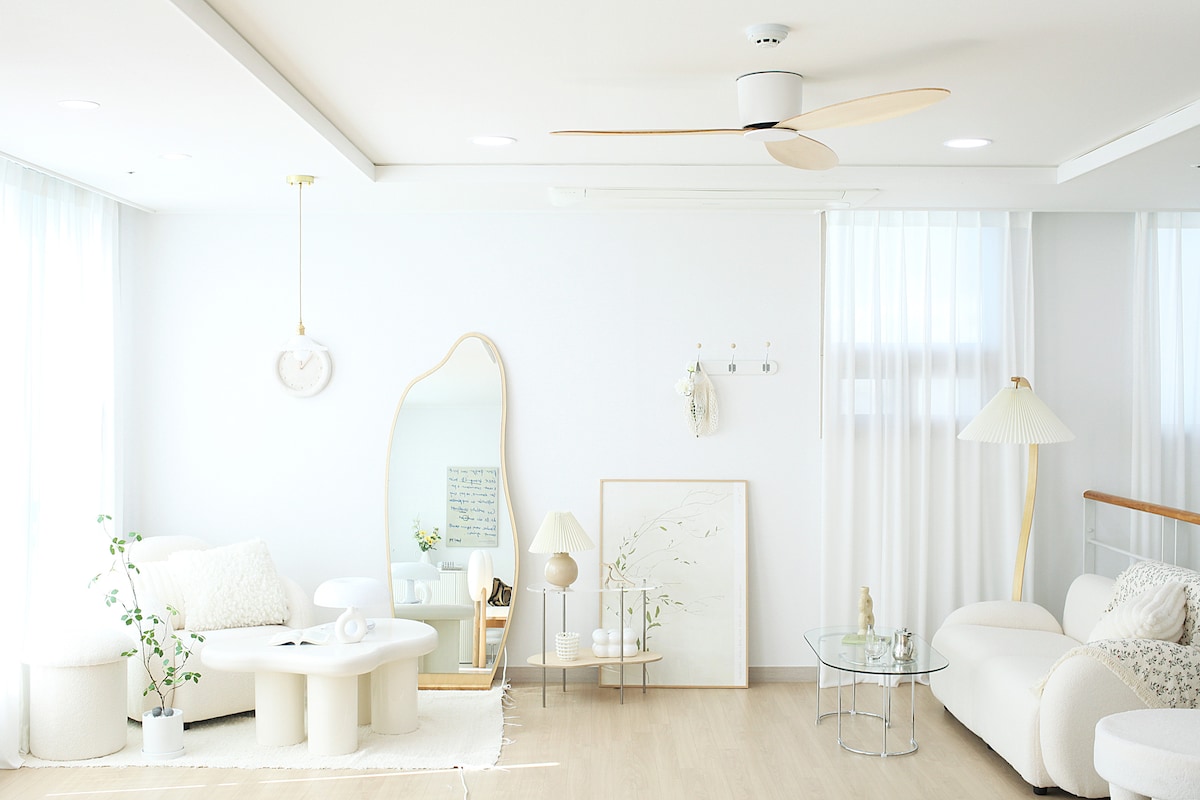 A bright living area features a neutral color palette with a spacious white sofa and a glass coffee table. A large mirror leans against the wall, complementing light wood flooring. Natural light streams in through sheer curtains, enhancing the open and airy ambiance.