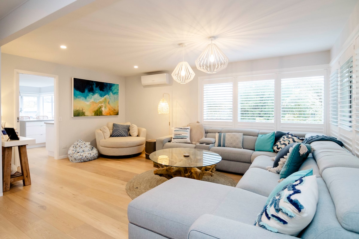 A bright living area is furnished with a spacious sectional couch and a circular glass coffee table. Natural light filters through the multiple windows adorned with white shutters, while decorative elements like paintings and cushions add subtle color and texture to the room.