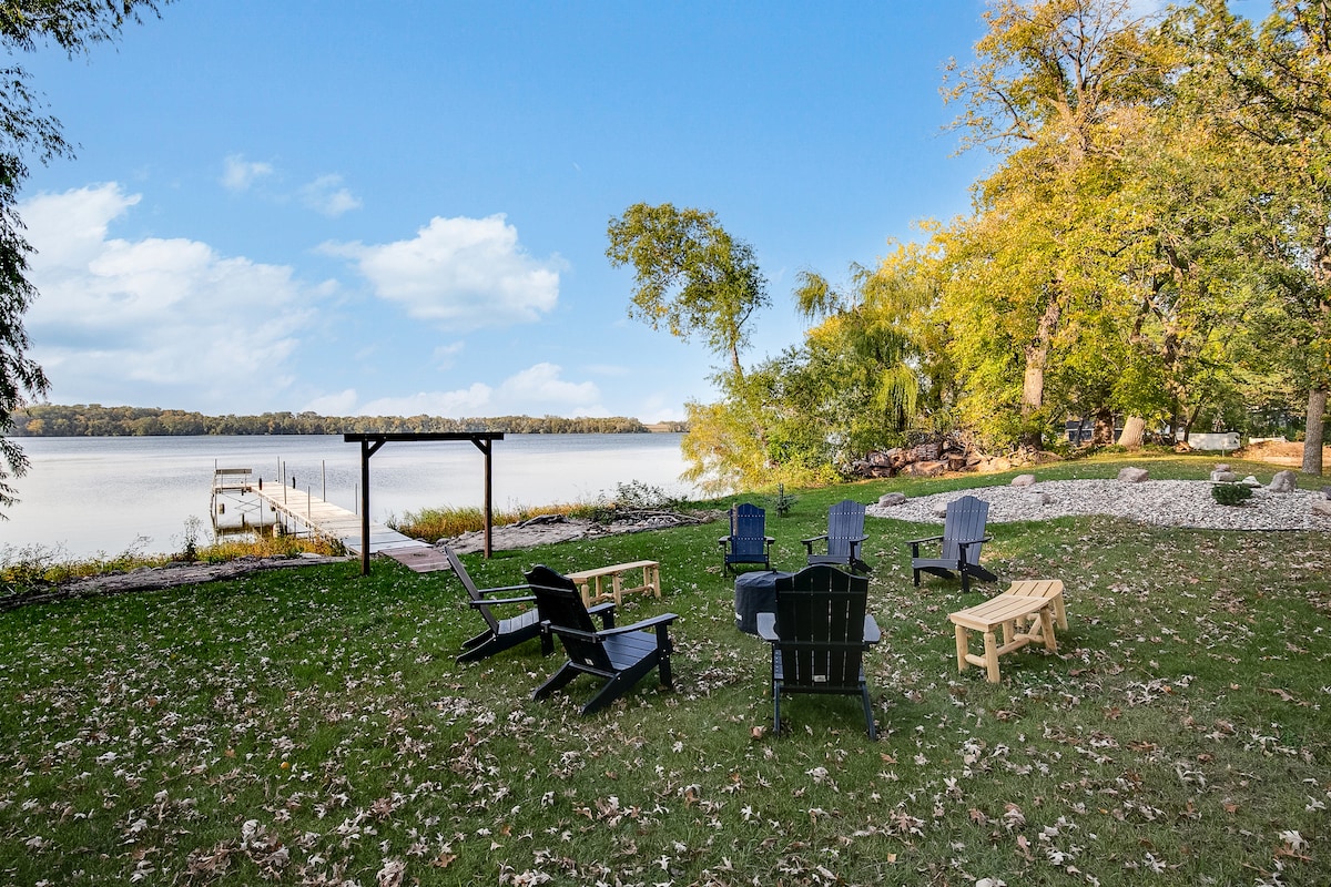 A spacious backyard features a well-maintained lawn surrounded by trees. Several Adirondack chairs are arranged around a fire pit, while a private dock extends into the lake, offering tranquil views of the water. The area is ideal for outdoor gatherings and relaxation.