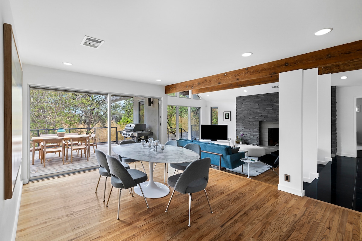 A bright and airy living space is showcased with large sliding doors that open to a deck and outdoor dining area. A modern dining table is surrounded by sleek chairs, while comfortable seating and a stone fireplace create a welcoming atmosphere. Natural light illuminates the space.