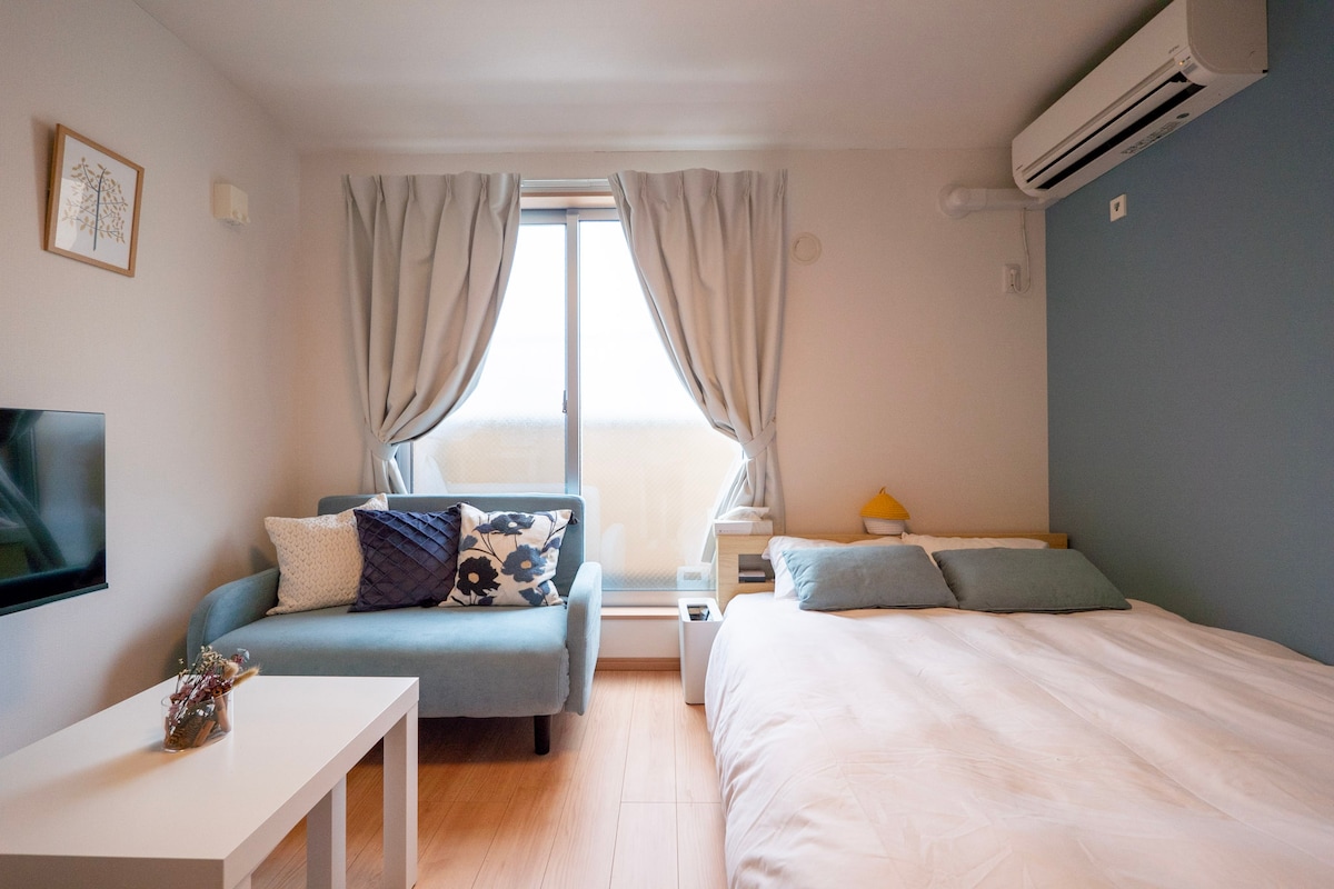 A bright room is presented, featuring a spacious bed and a blue sofa adorned with decorative cushions. Natural light enters through a large window, draped with sheer curtains. A small table is positioned beside the bed, enhancing the room's functionality.