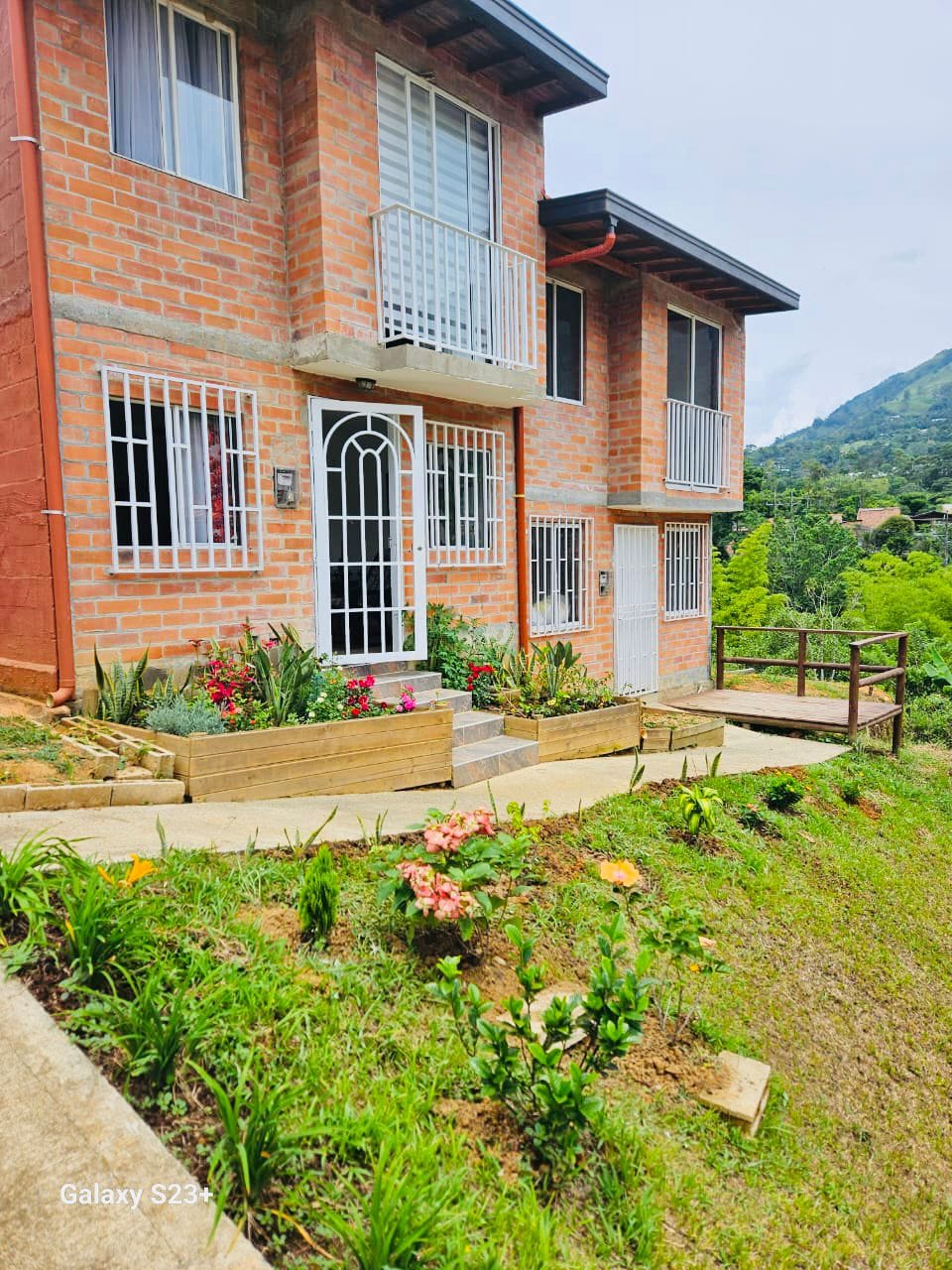 The brick two-story building is surrounded by vibrant flower beds and lush greenery. A pathway leads to the entrance, which features a decorative wrought iron door. Balconies on the upper level provide outdoor views of the landscape.