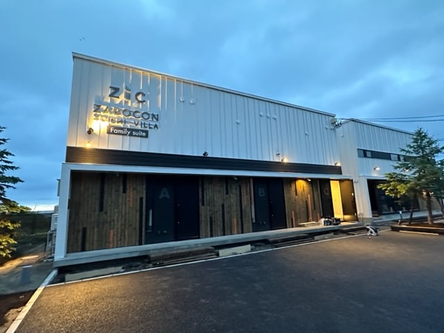 The exterior of the Family Suite building is depicted, featuring a modern design with a light-colored facade and wooden accents. Multiple doors are visible, labeled A, B, and C, indicating separate units. Evening lighting provides a soft glow, enhancing the welcoming atmosphere.