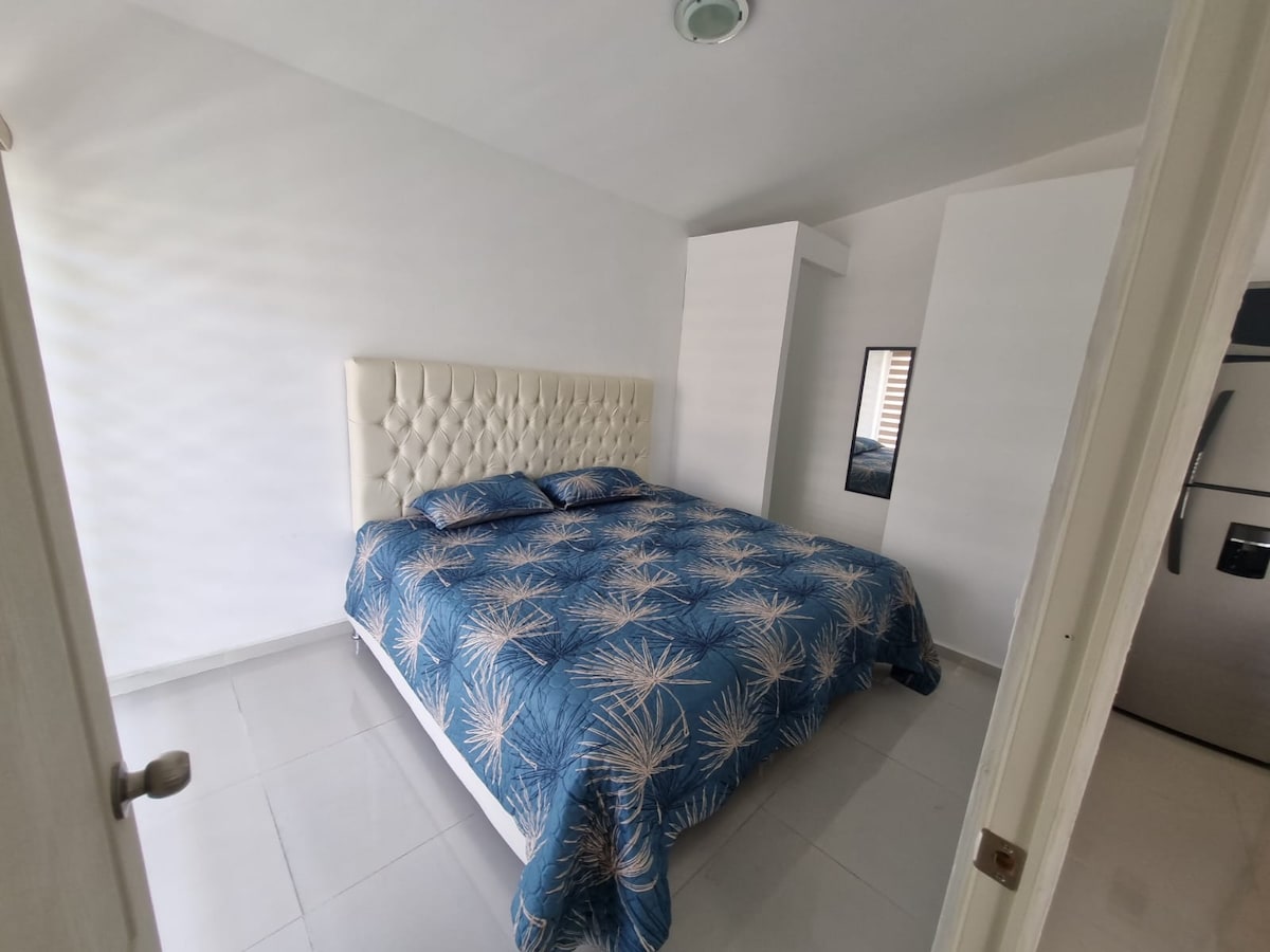 A bright bedroom features a spacious bed with a patterned blue comforter. Soft natural light enters through a nearby window, creating a calm atmosphere. A mirror is positioned on the wall opposite the bed, reflecting the clean, modern decor.
